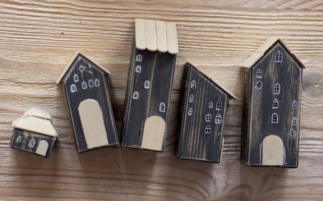 A series of five small, handcrafted wooden houses with black bodies and beige roofs, placed on a wooden surface. Each house is decorated with simple white window and door designs, giving a quaint village-like appearance.