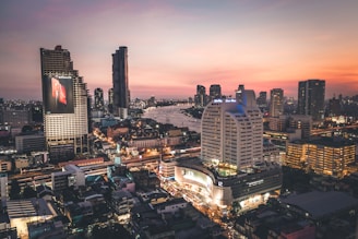 A vibrant cityscape at dusk with modern skyscrapers and high-rise buildings illuminated. There is a river running through the city, reflecting the warm hues of the sunset sky. Large billboards and advertisements can be seen on the buildings, with streets bustling with activity.