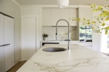 A modern kitchen with marble countertops and a sleek, chrome faucet in the center of an island. The cabinets are a light neutral color, providing a bright and clean aesthetic. A potted plant with yellow flowers adds a touch of nature to the space. In the background, large windows let in natural light, creating a vibrant and airy atmosphere.