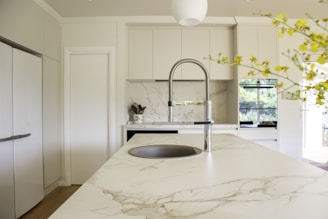 A modern kitchen with marble countertops and a sleek, chrome faucet in the center of an island. The cabinets are a light neutral color, providing a bright and clean aesthetic. A potted plant with yellow flowers adds a touch of nature to the space. In the background, large windows let in natural light, creating a vibrant and airy atmosphere.