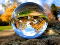 A crystal ball resting on a rock reflects an inverted scene of a vibrant autumn landscape, complete with colorful trees and a clear sky.