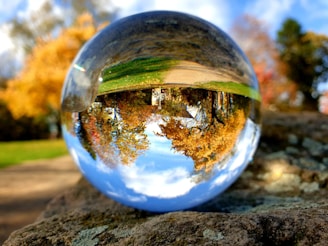 A crystal ball resting on a rock reflects an inverted scene of a vibrant autumn landscape, complete with colorful trees and a clear sky.