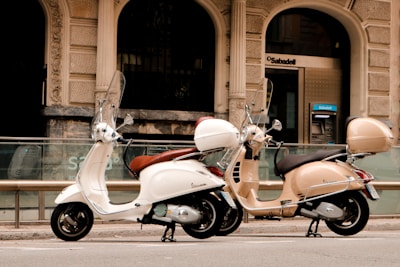 Two scooters are parked on a city street in front of a bank with ornate architecture. One scooter is white while the other is beige, both featuring windshields and storage compartments at the back.