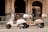 Two scooters are parked on a city street in front of a bank with ornate architecture. One scooter is white while the other is beige, both featuring windshields and storage compartments at the back.