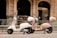 Two scooters are parked on a city street in front of a bank with ornate architecture. One scooter is white while the other is beige, both featuring windshields and storage compartments at the back.