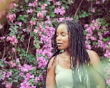 A woman with long braided hair stands in front of a lush backdrop of vibrant purple flowers and green foliage. She appears serene, with a peaceful expression and closed eyes. The natural setting creates a harmonious blend with her presence.