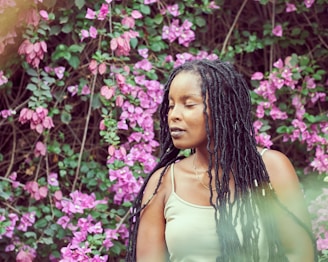 A woman with long braided hair stands in front of a lush backdrop of vibrant purple flowers and green foliage. She appears serene, with a peaceful expression and closed eyes. The natural setting creates a harmonious blend with her presence.