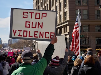 A large crowd of people participating in a protest, with one prominent sign reading 'Stop Gun Violence'. The protest is happening in an urban setting with tall buildings. An American flag is visible amongst the crowd.