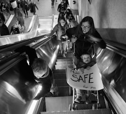 A woman and a child holding a sign reading 'Make it SAFE for me' are on an escalator in a crowded area. The child is in a stroller, and several people are ahead of them on the escalator. The setting appears to be indoors, with a tiled floor visible at the top of the escalator.