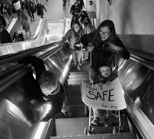 A woman and a child holding a sign reading 'Make it SAFE for me' are on an escalator in a crowded area. The child is in a stroller, and several people are ahead of them on the escalator. The setting appears to be indoors, with a tiled floor visible at the top of the escalator.