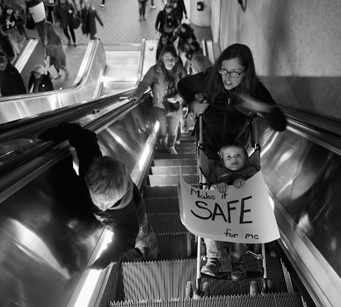 A woman and a child holding a sign reading 'Make it SAFE for me' are on an escalator in a crowded area. The child is in a stroller, and several people are ahead of them on the escalator. The setting appears to be indoors, with a tiled floor visible at the top of the escalator.