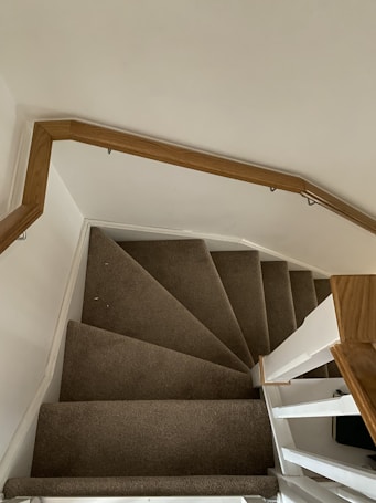 A top-down view of a carpeted spiral staircase with a wooden handrail. The steps are covered in a dark brown carpet, with the staircase structure painted white. The wall beside the staircase is painted light beige.
