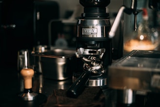 A close-up of a sophisticated espresso machine setup showing a coffee grinder and portafilter alongside other barista tools on a shiny countertop. The lighting is warm, highlighting the metallic surfaces and creating a cozy atmosphere.