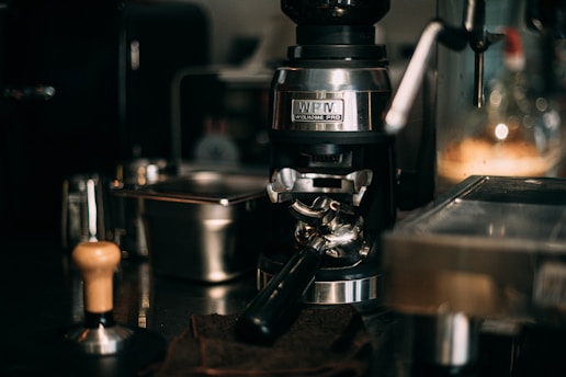 A close-up of a sophisticated espresso machine setup showing a coffee grinder and portafilter alongside other barista tools on a shiny countertop. The lighting is warm, highlighting the metallic surfaces and creating a cozy atmosphere.