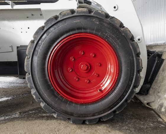A close-up view of a large, rugged tire with a bright red hubcap mounted on a heavy-duty vehicle. The tire sidewall shows the brand name 'GALAXY' embossed on it. The surrounding area includes parts of the vehicle’s white frame and a concrete floor.