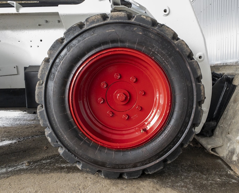 A close-up view of a large, rugged tire with a bright red hubcap mounted on a heavy-duty vehicle. The tire sidewall shows the brand name 'GALAXY' embossed on it. The surrounding area includes parts of the vehicle&rsquo;s white frame and a concrete floor.