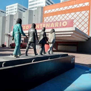 Four statues of people walking in a line are positioned on a platform in front of a modern building with the sign 'CENTENARIO'. The statues are stylized, with each figure wearing different clothing, and they are facing away from the viewer. The architecture of the building features vertical lines and a prominent orange grid design.