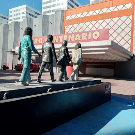 Four statues of people walking in a line are positioned on a platform in front of a modern building with the sign 'CENTENARIO'. The statues are stylized, with each figure wearing different clothing, and they are facing away from the viewer. The architecture of the building features vertical lines and a prominent orange grid design.