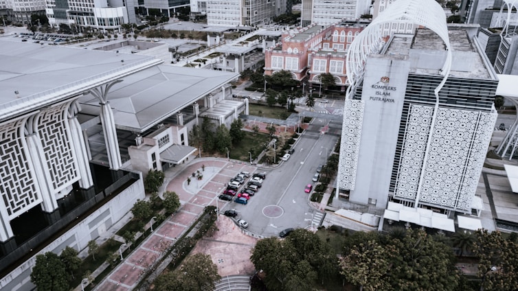 An aerial view of a modern architectural complex featuring large buildings with detailed geometric patterns. The scene includes a city street with several parked cars and surrounding greenery. A prominent structure has a sign reading 'Kompleks Islam Putrajaya'. The design combines contemporary and traditional Islamic architectural elements.
