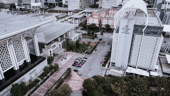 An aerial view of a modern architectural complex featuring large buildings with detailed geometric patterns. The scene includes a city street with several parked cars and surrounding greenery. A prominent structure has a sign reading 'Kompleks Islam Putrajaya'. The design combines contemporary and traditional Islamic architectural elements.