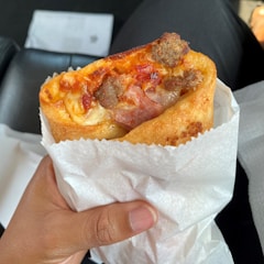 A hand holding a folded, paper-wrapped pizza cone filled with melted cheese, pieces of meat, and tomato sauce. The background appears to be a car's interior with a partial view of black seats and a white envelope.
