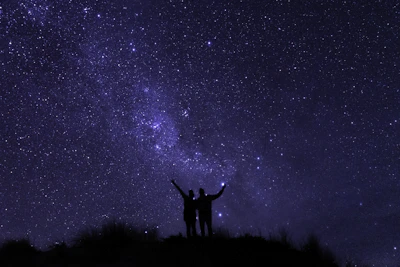 A silhouette of two figures reaching out to each other beneath a starry sky filled with swirling constellations.