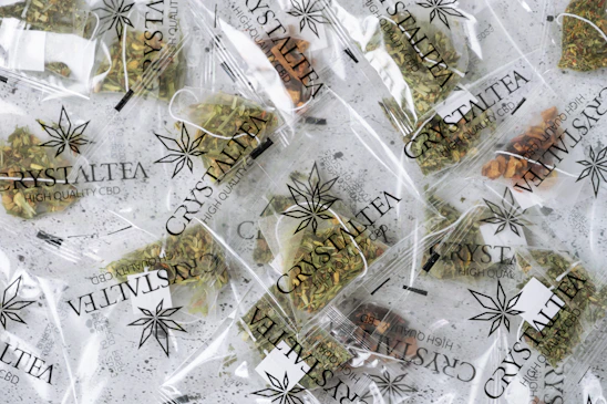 Multiple small transparent bags filled with dried herbal tea labeled 'CrystalTea High Quality CBD'. The bags display a leaf logo, and the herbs inside vary in color with some green and brown hues visible.