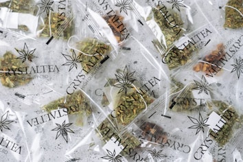 Multiple small transparent bags filled with dried herbal tea labeled 'CrystalTea High Quality CBD'. The bags display a leaf logo, and the herbs inside vary in color with some green and brown hues visible.