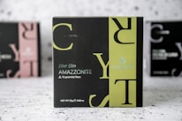 A product box with the label 'CRYSTALTEA' is prominently displayed in the center with a black and olive green design. The text includes 'Elixir Slim AMAZZONITE' along with a note about '10 pyramidal filters'. The background features a blurred image of similar product boxes.