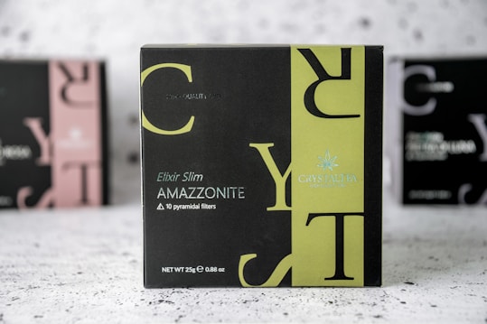 A product box with the label 'CRYSTALTEA' is prominently displayed in the center with a black and olive green design. The text includes 'Elixir Slim AMAZZONITE' along with a note about '10 pyramidal filters'. The background features a blurred image of similar product boxes.