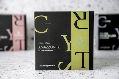 A product box with the label 'CRYSTALTEA' is prominently displayed in the center with a black and olive green design. The text includes 'Elixir Slim AMAZZONITE' along with a note about '10 pyramidal filters'. The background features a blurred image of similar product boxes.