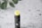 A tube of lip balm with a black container and a yellow balm protruding from the top. The container has a minimalist design with a small logo. It's placed on a textured white surface with blurred green leaves in the background.
