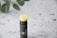 A tube of lip balm with a black container and a yellow balm protruding from the top. The container has a minimalist design with a small logo. It's placed on a textured white surface with blurred green leaves in the background.