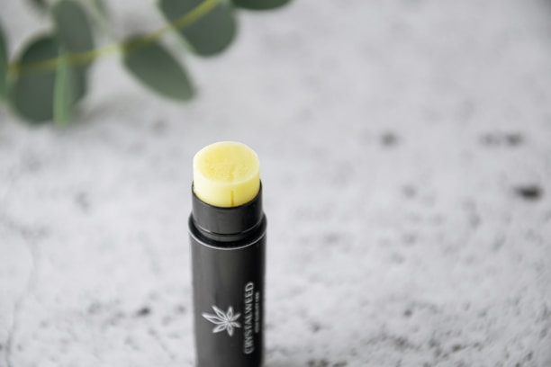 A tube of lip balm with a black container and a yellow balm protruding from the top. The container has a minimalist design with a small logo. It's placed on a textured white surface with blurred green leaves in the background.