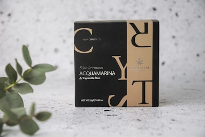 A box labeled 'Elixir Immuno Acquamarina' featuring a sleek, modern design with contrasting black and beige colors. The text includes details about the content, highlighting '10 pyramidal filters' and 'Net WT 25g'. The packaging also includes branding logo and text. A eucalyptus branch is positioned on the left, adding a natural element, set against a textured, speckled background.