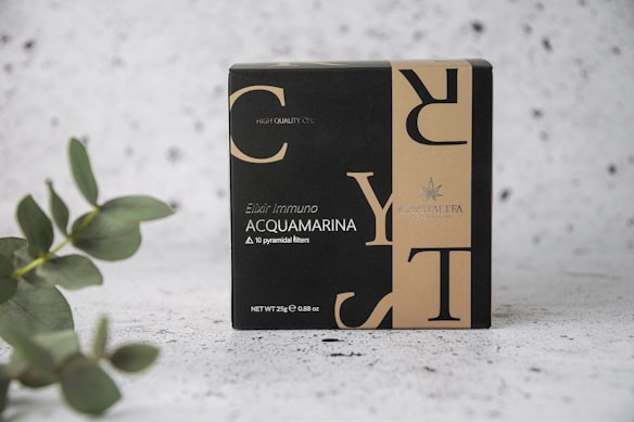 A box labeled 'Elixir Immuno Acquamarina' featuring a sleek, modern design with contrasting black and beige colors. The text includes details about the content, highlighting '10 pyramidal filters' and 'Net WT 25g'. The packaging also includes branding logo and text. A eucalyptus branch is positioned on the left, adding a natural element, set against a textured, speckled background.