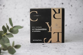 A box labeled 'Elixir Immuno Acquamarina' featuring a sleek, modern design with contrasting black and beige colors. The text includes details about the content, highlighting '10 pyramidal filters' and 'Net WT 25g'. The packaging also includes branding logo and text. A eucalyptus branch is positioned on the left, adding a natural element, set against a textured, speckled background.