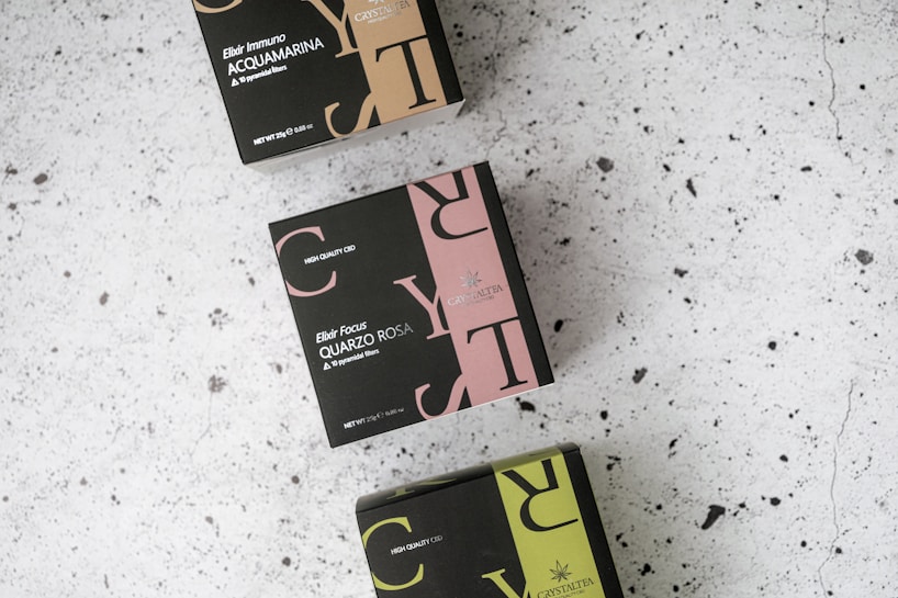 Three boxes with elegant, modern designs arranged on a speckled, light-colored surface. The boxes feature bold typography in colors such as beige, pink, and green, with text indicating different varieties of tea blends, including Acquamarina and Quarzo Rosa. Each box is labeled with 'High Quality CBD' and shows details like net weight.
