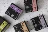 Multiple boxes of herbal tea products are arranged on a speckled surface. Each box is uniquely colored, featuring a distinctive design with bold letters and branding, including product names like 'Elixir Detox AGATA' and 'Elixir Relax PIETRA DI LUNA'. The colors range from purples and greens to browns, with prominent logos displayed on each box.