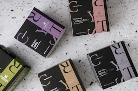 Multiple boxes of herbal tea products are arranged on a speckled surface. Each box is uniquely colored, featuring a distinctive design with bold letters and branding, including product names like 'Elixir Detox AGATA' and 'Elixir Relax PIETRA DI LUNA'. The colors range from purples and greens to browns, with prominent logos displayed on each box.