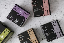 Multiple boxes of herbal tea products are arranged on a speckled surface. Each box is uniquely colored, featuring a distinctive design with bold letters and branding, including product names like 'Elixir Detox AGATA' and 'Elixir Relax PIETRA DI LUNA'. The colors range from purples and greens to browns, with prominent logos displayed on each box.