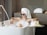 A person relaxes in a stylish freestanding bathtub filled with bubbles, wearing sunglasses and a towel wrapped around their head. The bathroom has a modern feel, with a sleek faucet and a collection of colorful bottles on a small table nearby.