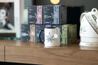 A wooden table holds a white mug with a black leaf design, a tea bag tag hanging from its rim. Nearby is a teapot with similar black geometric patterns. Several boxes with varying colors, possibly tea or coffee packages, are arranged in the background. A blurred photo frame can be seen towards the left.