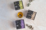 Three square boxes labeled with herbal tea names lie on a light textured surface. A glass cup filled with tea, containing a tea bag, is placed near the center. Two eucalyptus branches extend into the scene from the edges.