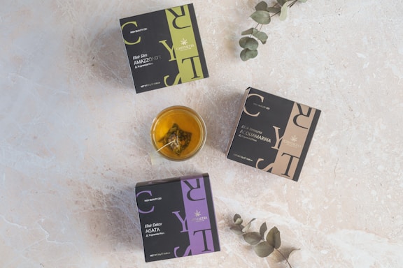 Three square boxes labeled with herbal tea names lie on a light textured surface. A glass cup filled with tea, containing a tea bag, is placed near the center. Two eucalyptus branches extend into the scene from the edges.