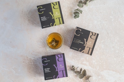 Three square boxes labeled with herbal tea names lie on a light textured surface. A glass cup filled with tea, containing a tea bag, is placed near the center. Two eucalyptus branches extend into the scene from the edges.