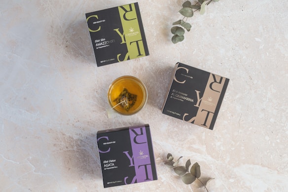 Three square boxes labeled with herbal tea names lie on a light textured surface. A glass cup filled with tea, containing a tea bag, is placed near the center. Two eucalyptus branches extend into the scene from the edges.