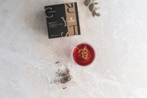 A marble surface displays a black and gold box labeled 'Crystal Tea High Quality CBD Elixir Immuno Acquamarina'. Next to it, there's a transparent container holding a red liquid with a tea bag inside visible. A small plant with green leaves is placed near the objects.
