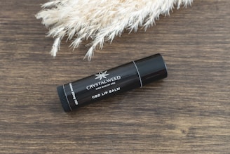 A black tube of CBD lip balm is placed on a wooden surface next to a bunch of light-colored, fluffy pampas grass. The lip balm tube is labeled 'CRYSTALWEED' and 'CBD LIP BALM' along with the dosage information.