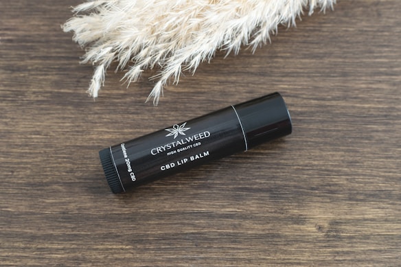 A black tube of CBD lip balm is placed on a wooden surface next to a bunch of light-colored, fluffy pampas grass. The lip balm tube is labeled 'CRYSTALWEED' and 'CBD LIP BALM' along with the dosage information.